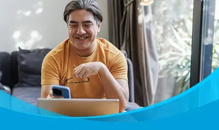 A smiling man in an orange shirt checks his phone while sitting at a laptop.